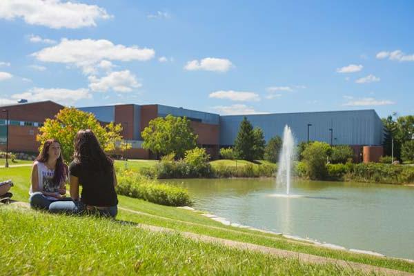 Two females students sitting on the lawn outside the student center in Spring near a flowing fountain. 